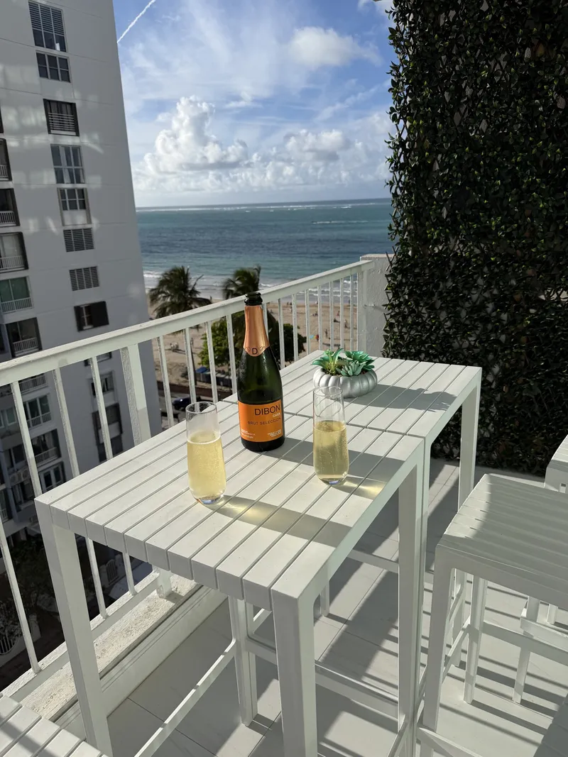 A bottle of cava and two poured glasses on the Terra Noir balcony table with Isla Verde Beach beyond the railing